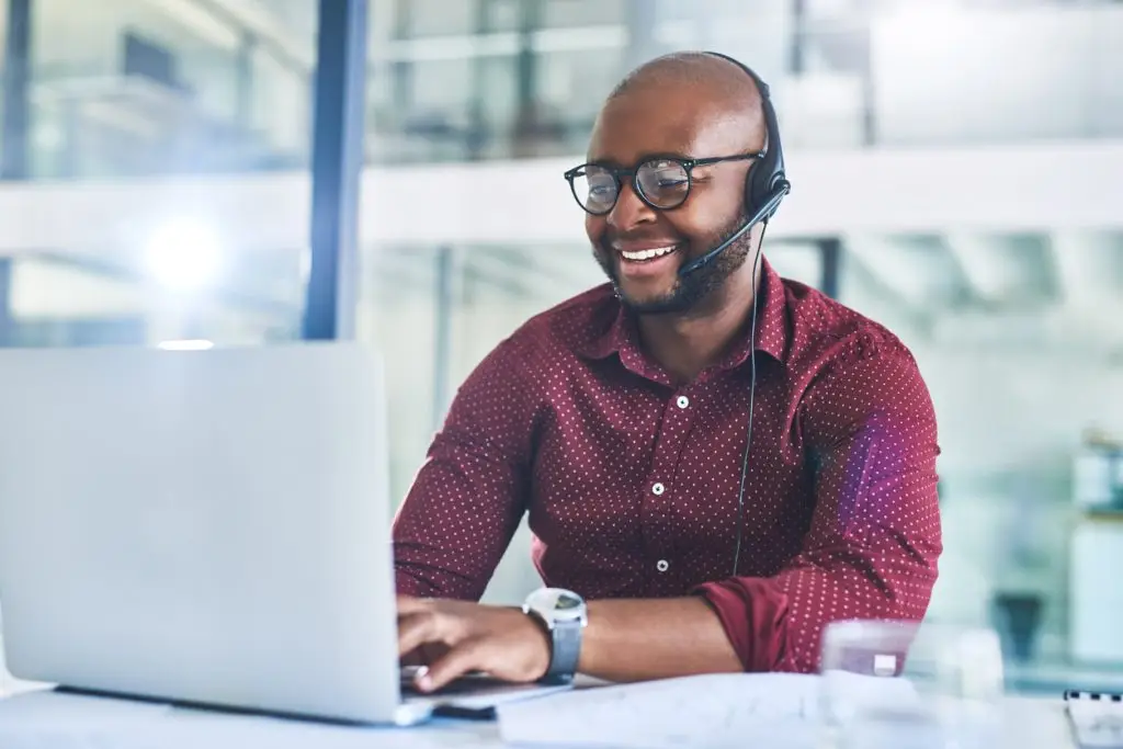 A smiling man wearing a headset working on a laptop in an office setting.