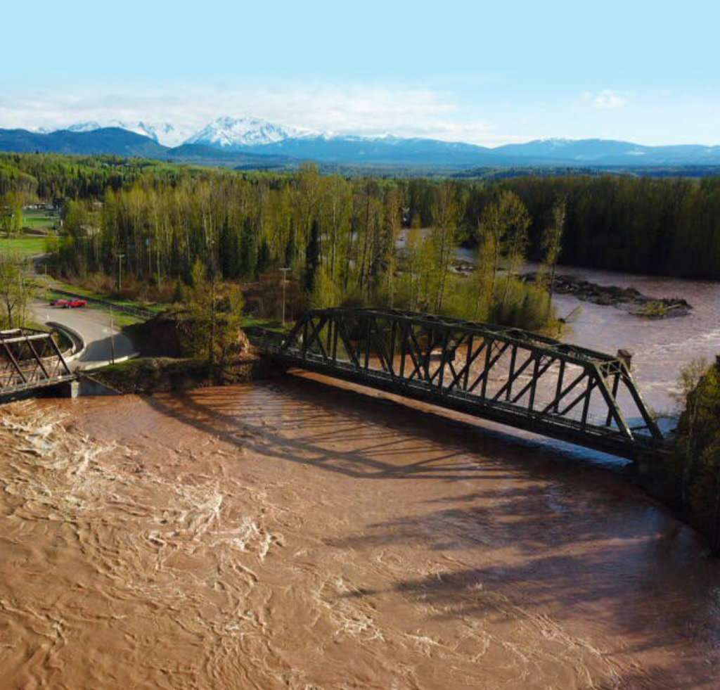Aerial view of a truss bridge over a muddy river with mountains in the background.