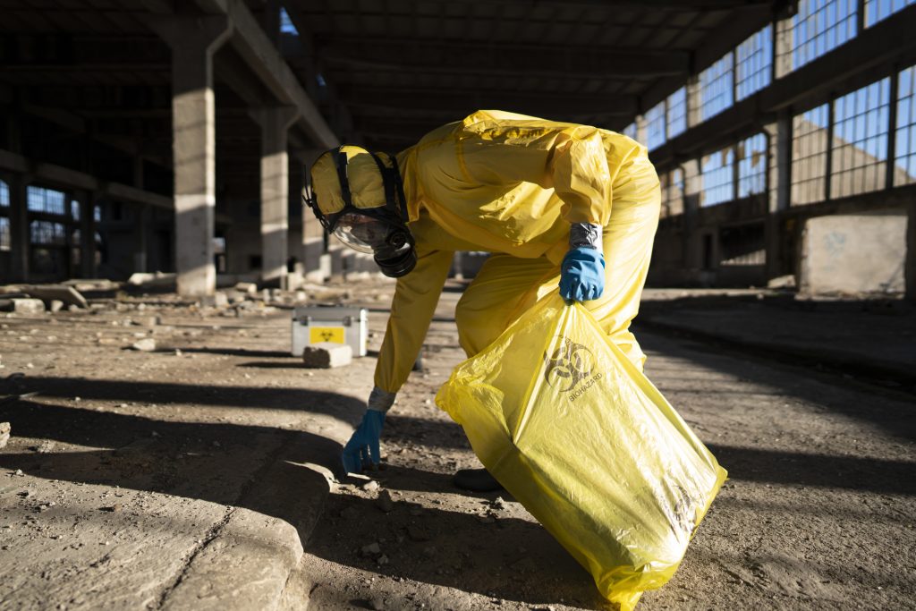 A person in a yellow protective suit picking up a bag of hazardous waste.