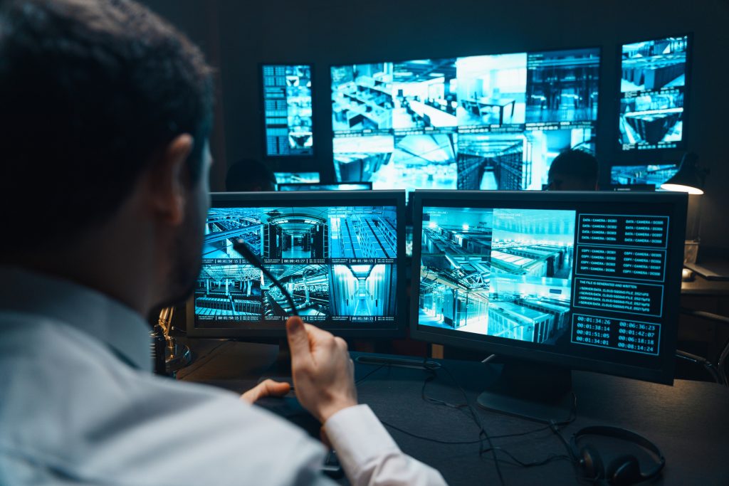 A police officer sitting at a desk with several surveillance monitors in front of him.
