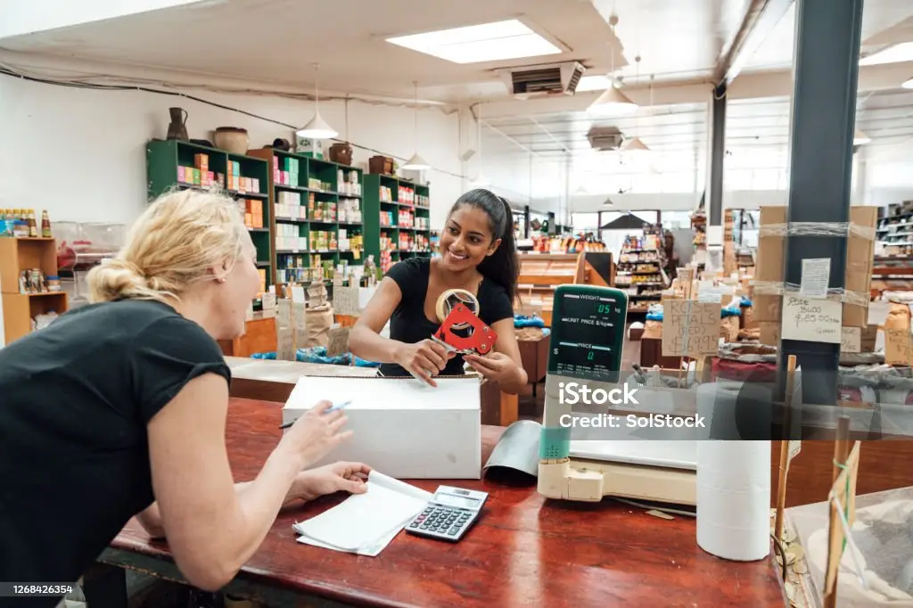 A smiling cashier assisting a customer at checkout in a store with shelves of goods in the background.