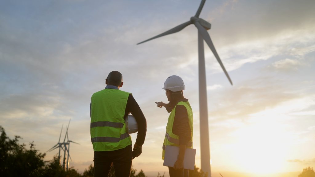 Two workers in safety vests and helmets stand near wind turbines at sunset, discussing or inspecting the site.