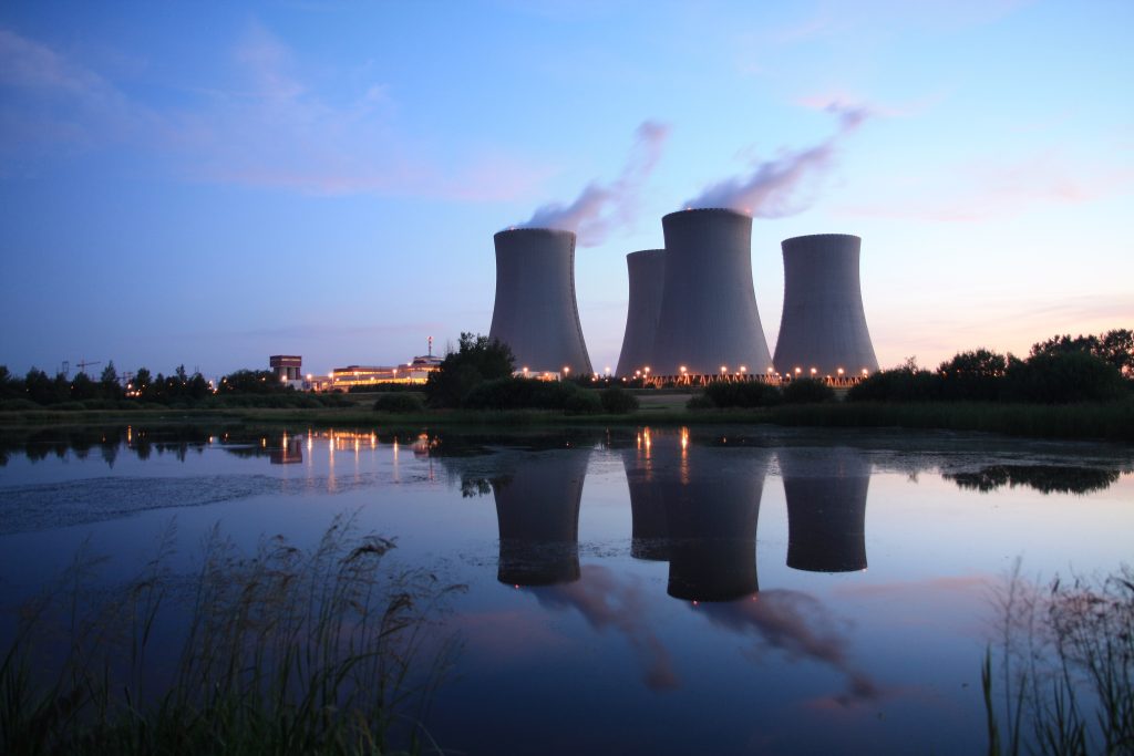 Cooling towers of a nuclear power plant release steam at dusk, with their reflection visible in a calm body of water in the foreground.