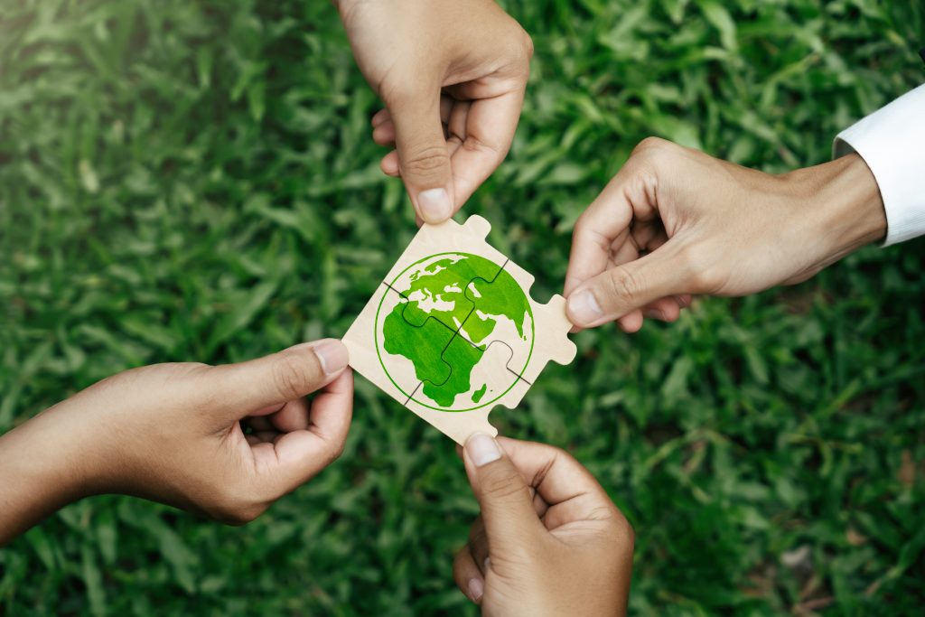 Four hands hold pieces of a jigsaw puzzle with a green Earth illustration, assembling it together over a background of green grass.