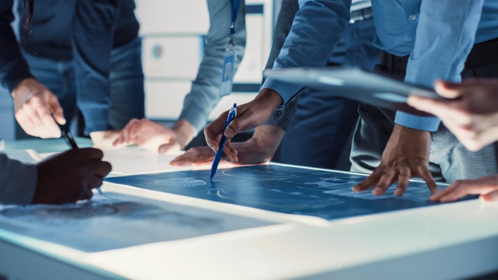 A group of professionals in an office setting review technical blueprints displayed on a glowing light table.