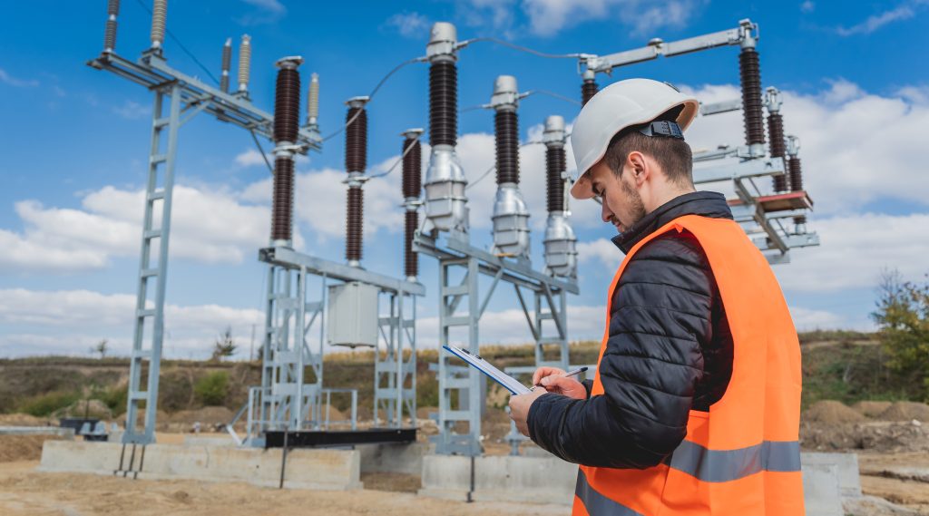 Engineer in a hard hat and safety vest writing on a clipboard in front of an electrical substation.