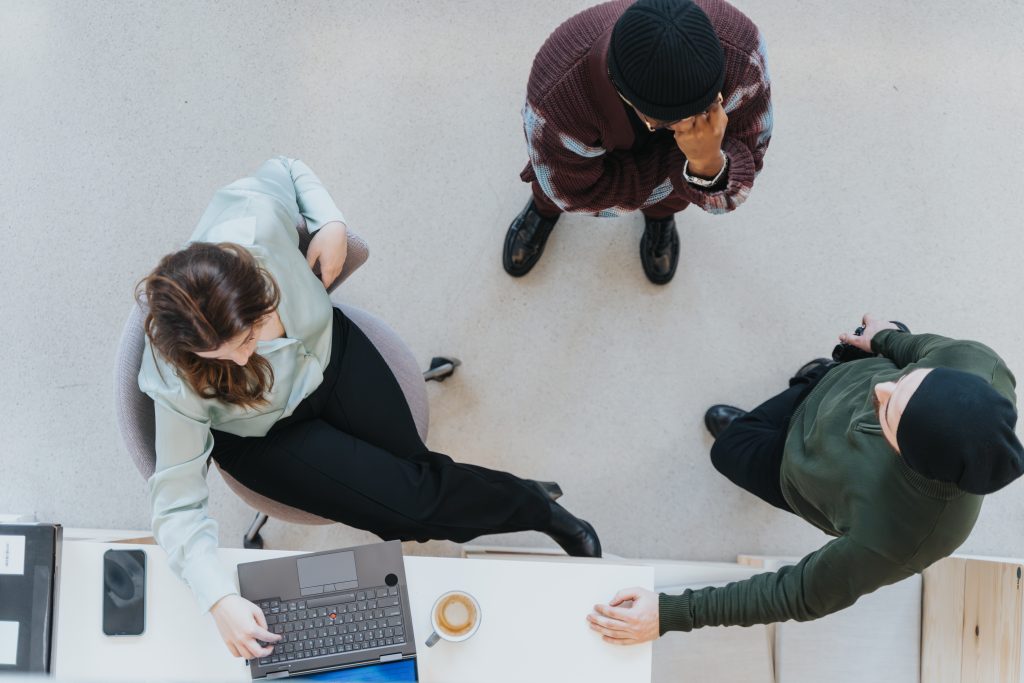 Three people gathered around a small table; one person sits at a laptop while two others stand nearby.
